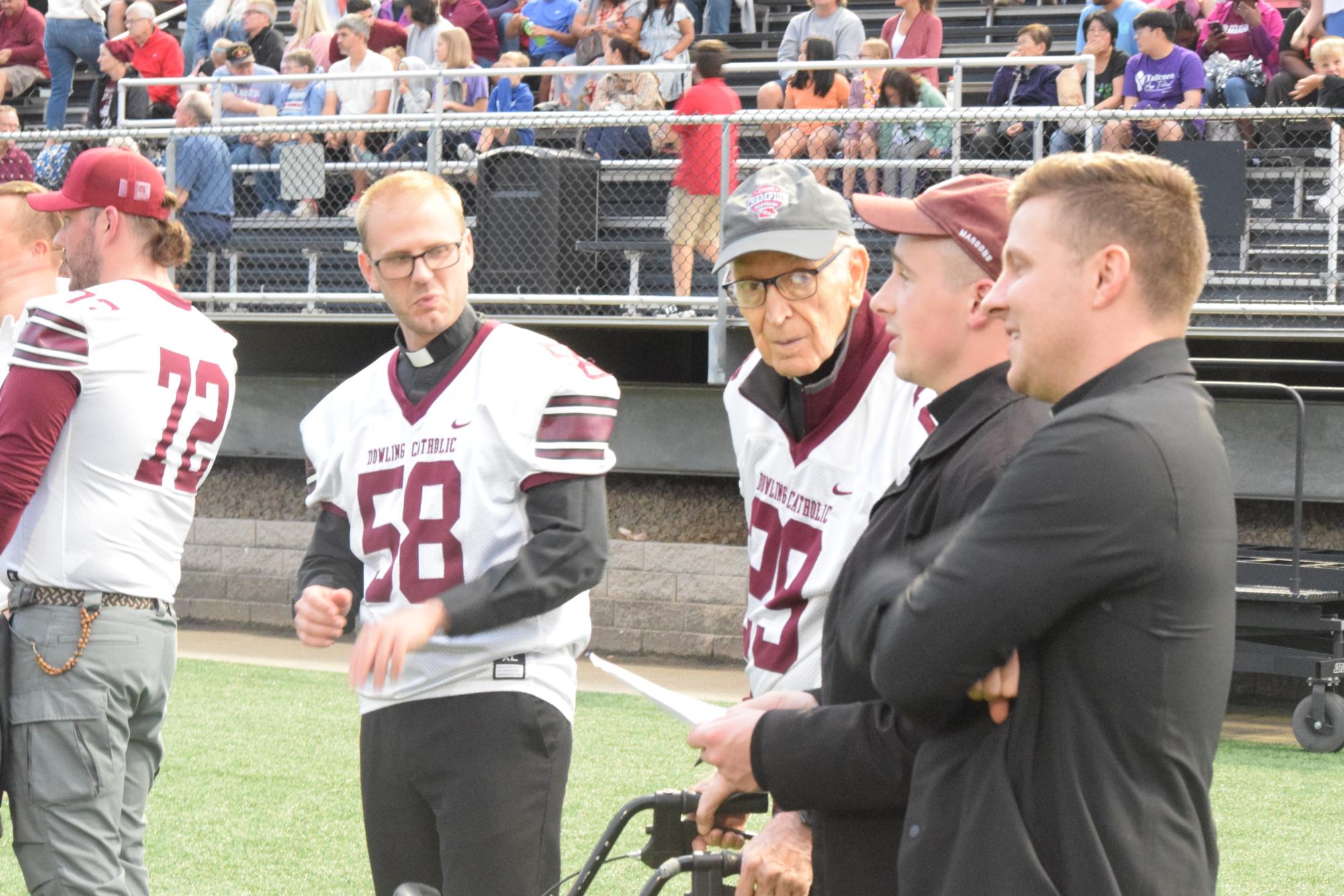 Father Andrew, Father Acrea, Father Downey, and Father Flood stand on the sideline at a football game.