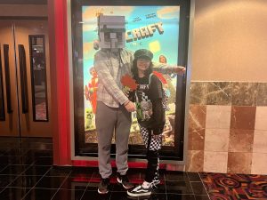 Brody Eberhart (left) and Becca Davila-Gay (right) posing in front of "A Minecraft Movie" poster